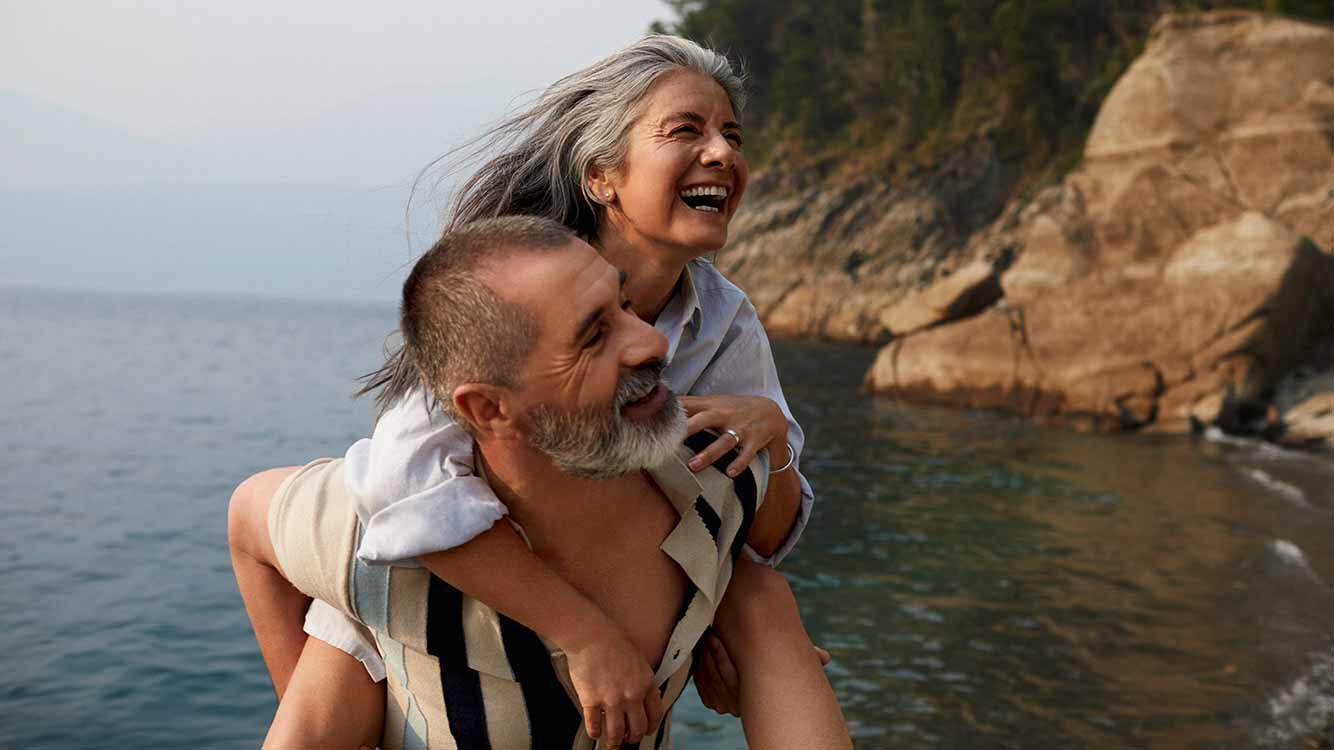 Un homme d'un certain &acirc;ge porte une femme d'un certain &acirc;ge sur son dos, sur la plage. En arri&egrave;re-plan, des falaises. Tous deux ont l'air tr&egrave;s heureux.